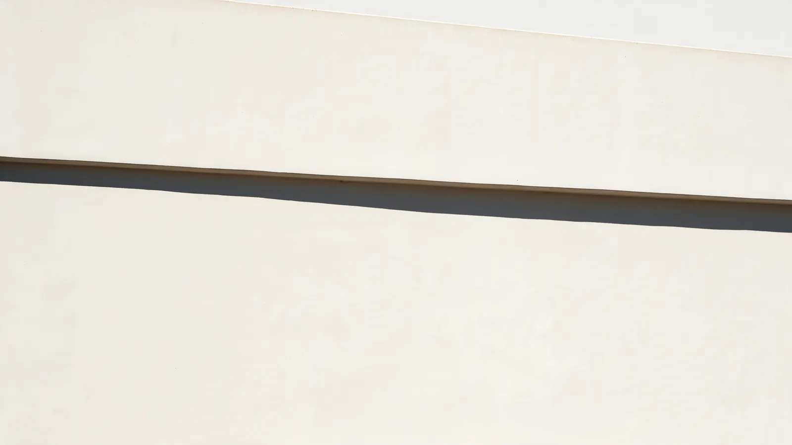 A long warm-white concrete wall crossed by one perfectly straight shadow line.
