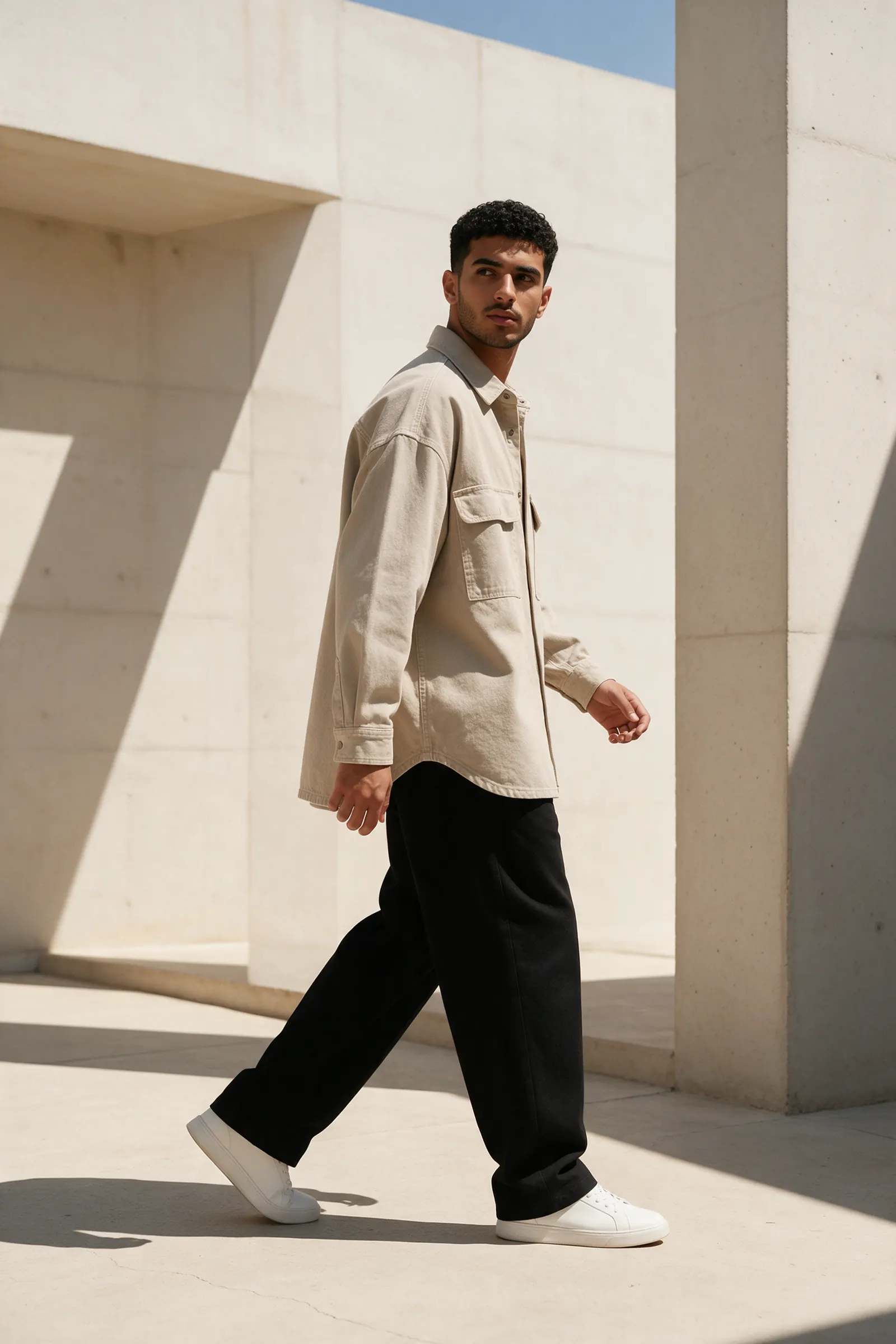 A young man in a stone overshirt and black trousers walks past bright concrete architecture.