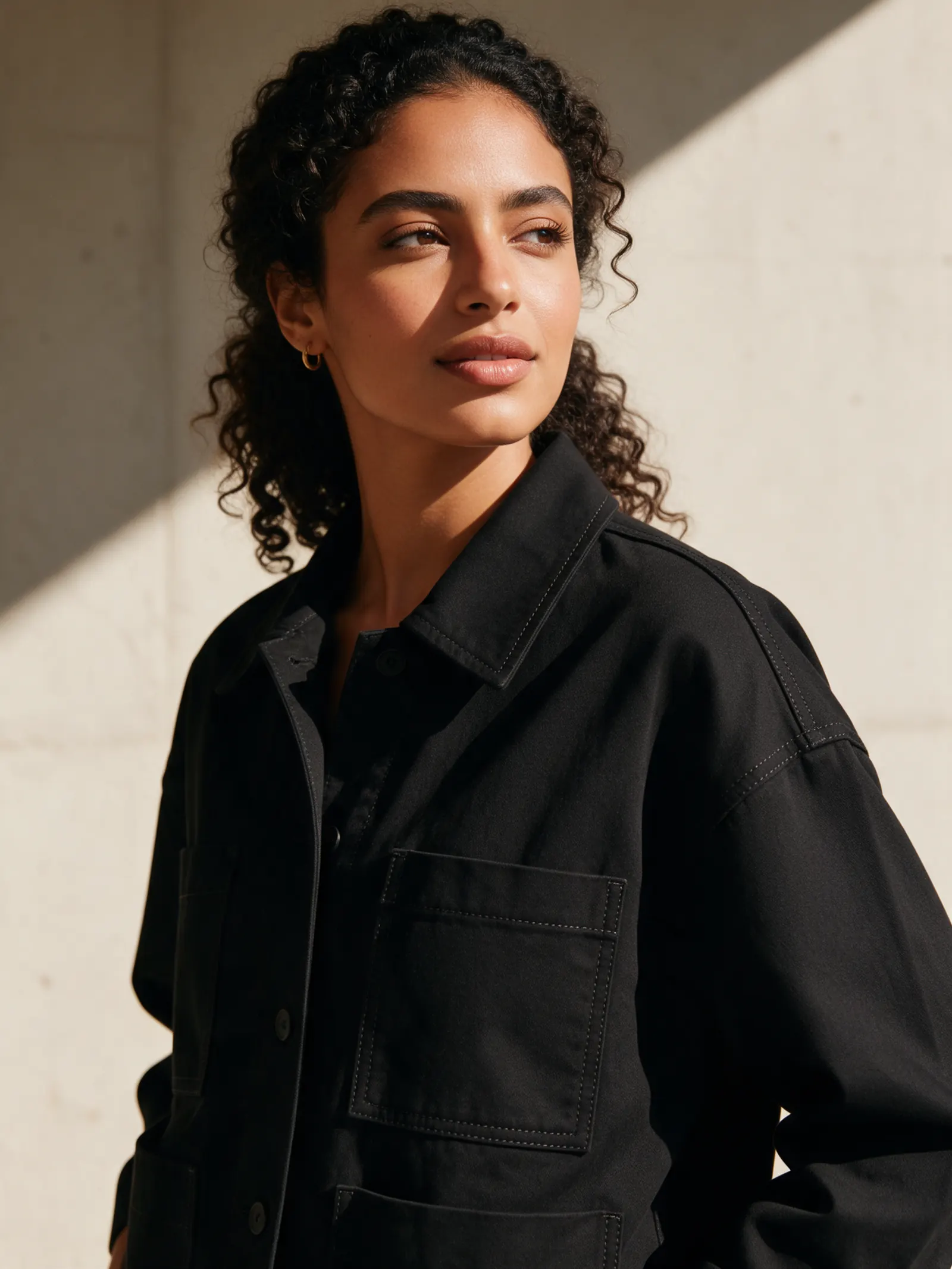 Portrait of a young woman in a black boxy jacket against a sunlit concrete wall.