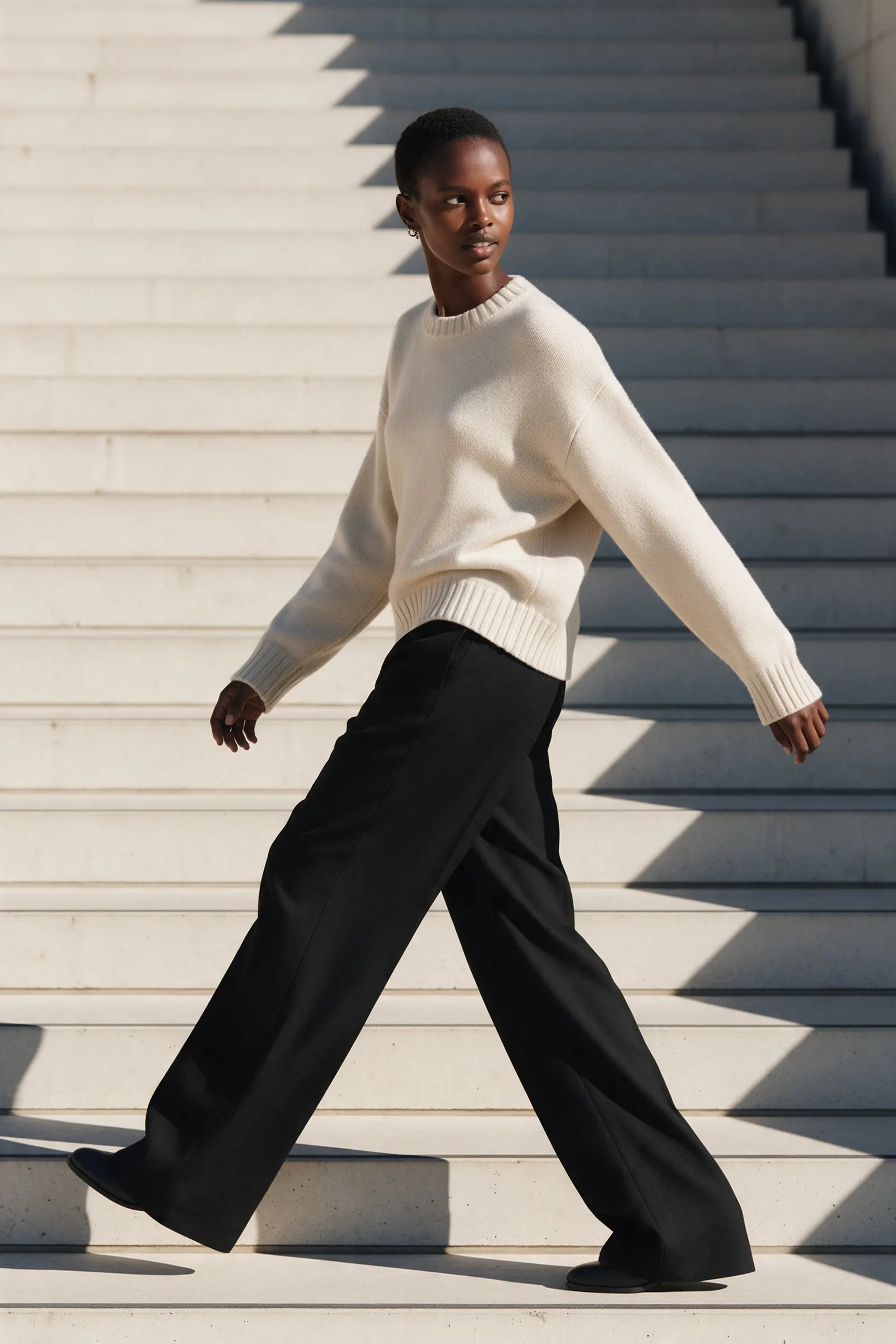 A person in a cream knit and black trousers walks up wide concrete steps.